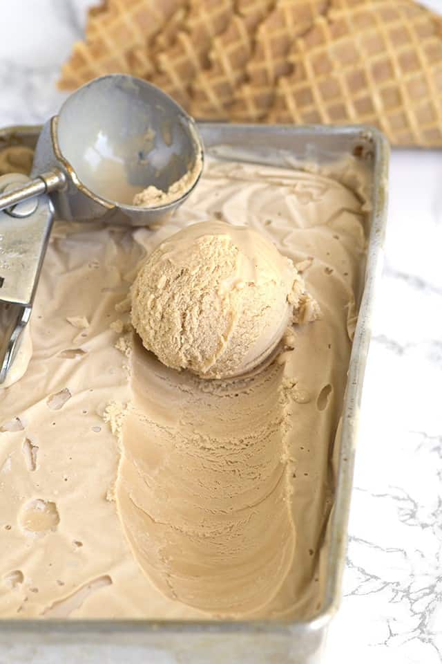 scoop of ice cream on top of the remaining ice cream with an ice cream scoop sitting next to it and waffle cones behind it